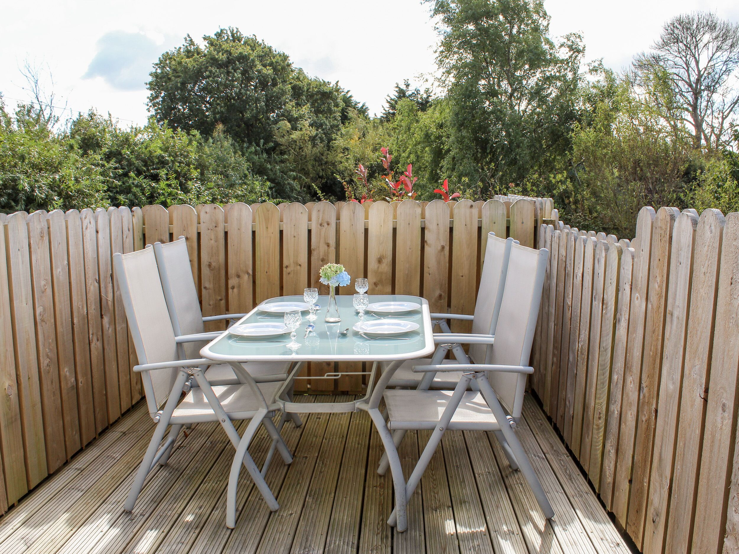Terraza de madera con una mesa de vidrio dispuesta para cuatro personas, rodeada de sillas blancas.