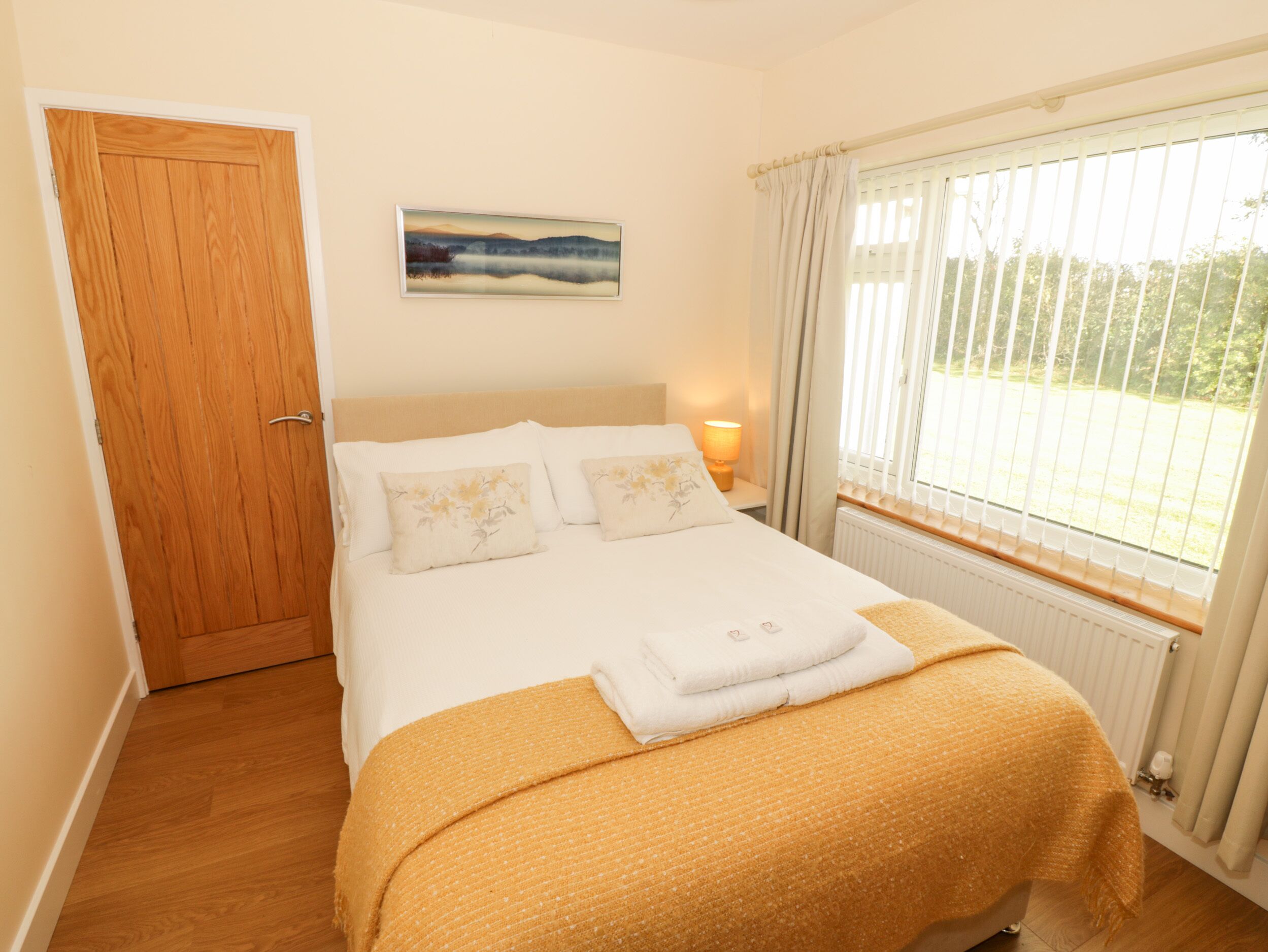 A neatly made bed with a white duvet and two pillows, a wooden door, a painting on the wall, and a window with curtains.