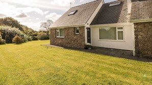 A stone house with a sloped roof, a small attached garage, and a well-maintained lawn. - Llain Bach (Llangefni)