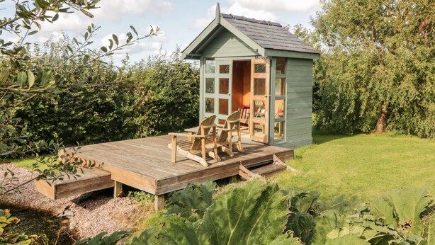 A small wooden cabin with a deck and two chairs, surrounded by greenery. - Llain Bach (Llangefni)
