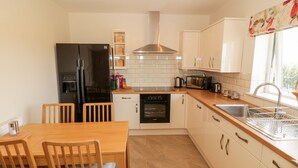 A modern kitchen with a black refrigerator, white cabinets, a wooden dining table with chairs, and a window with a floral curtain. - Llain Bach (Llangefni)