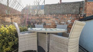 A patio with wicker furniture and a glass table, surrounded by a brick wall and a hedge. - Chapel Cottage (Filey)