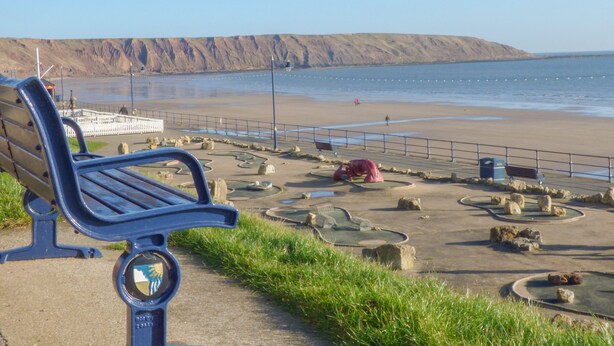 A bench overlooking a beach with a rocky shoreline and a distant mountain. - Chapel Cottage (Filey)