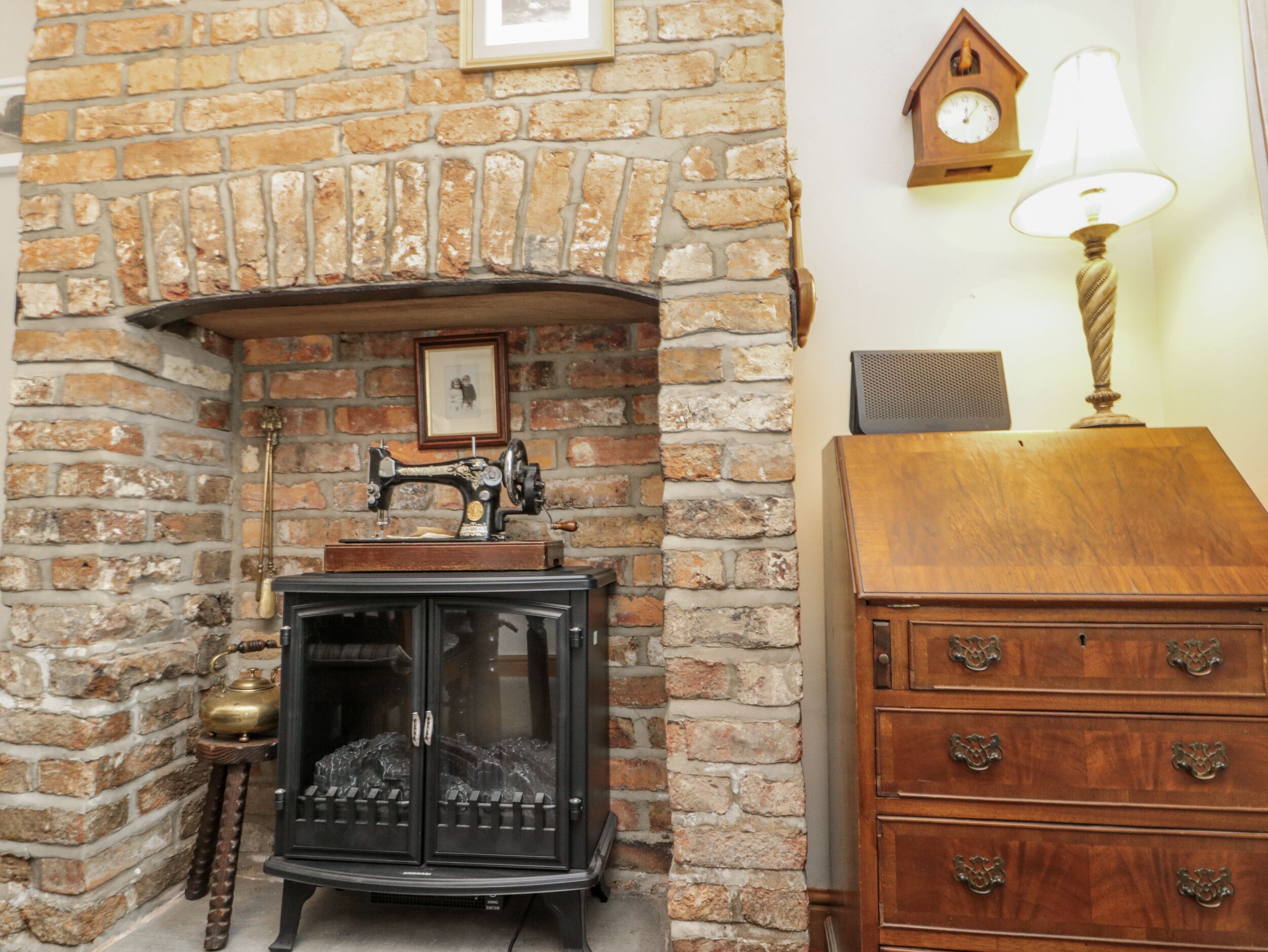 A room with a brick fireplace, a vintage sewing machine, a wooden dresser, and a wall clock.