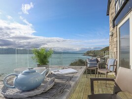 A terrace with a wicker table set with a teapot, cups, and a potted plant, overlooking a body of water and a mountainous landscape.