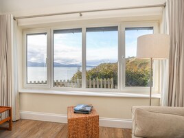 A room with a large window overlooking a body of water and hills, a wicker stool, a lamp, and a sofa.