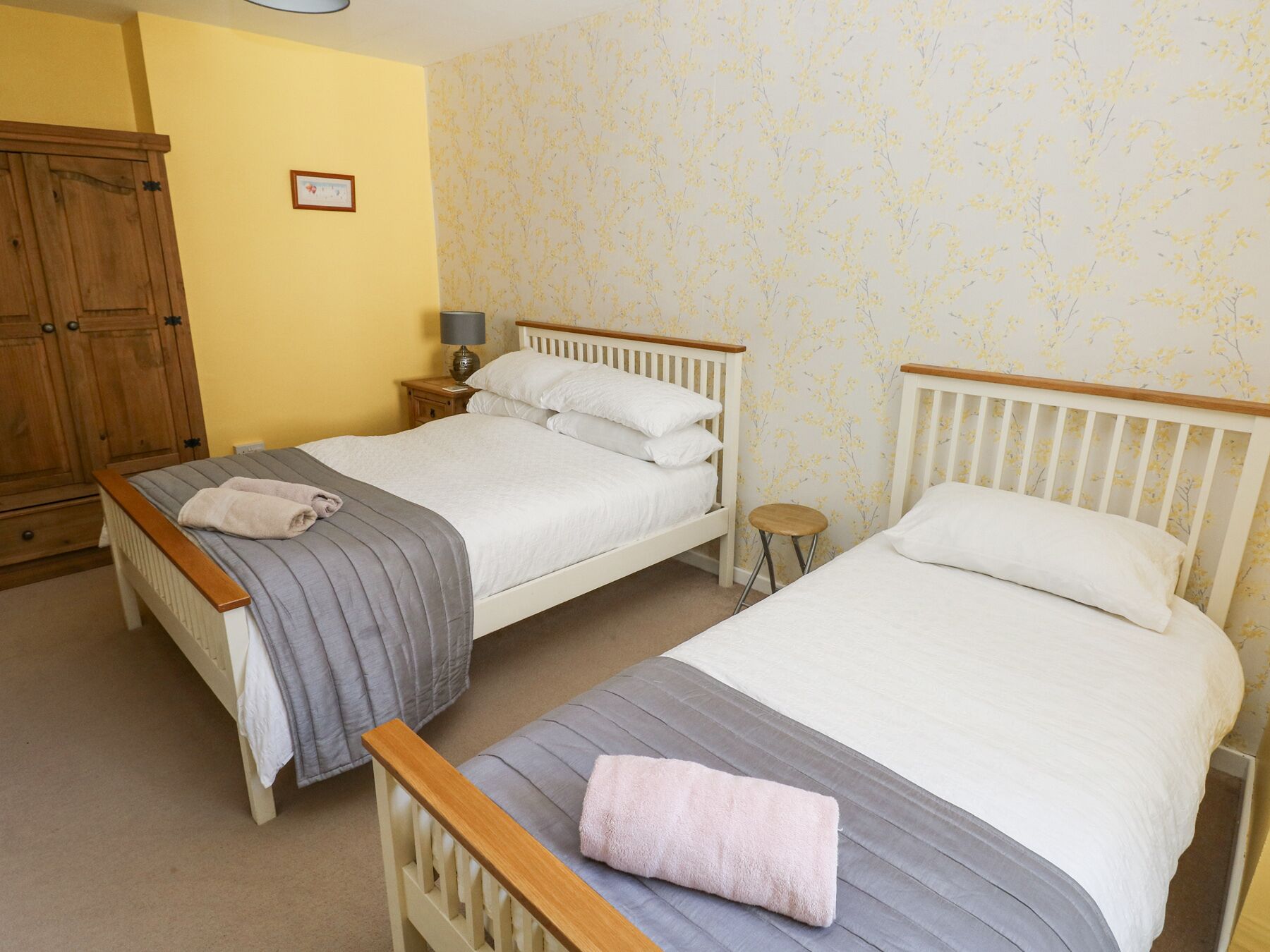 Une chambre avec deux lits simples, une armoire en bois, une petite table de chevet avec une lampe et un mur recouvert d’un papier peint à motifs floraux.