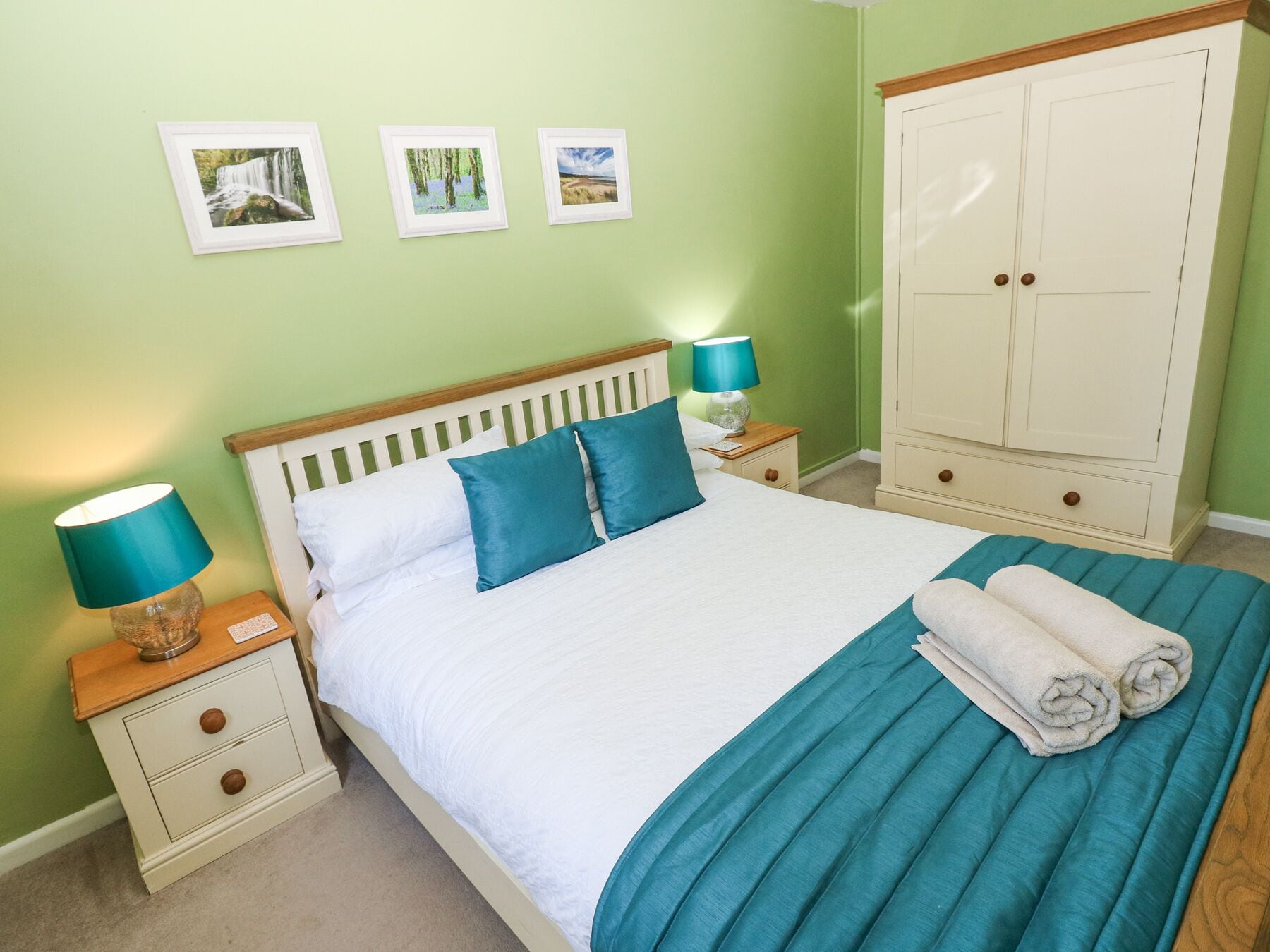 A neatly made bed with a blue blanket and pillows, a nightstand with a lamp, and a wardrobe in a room with green walls.