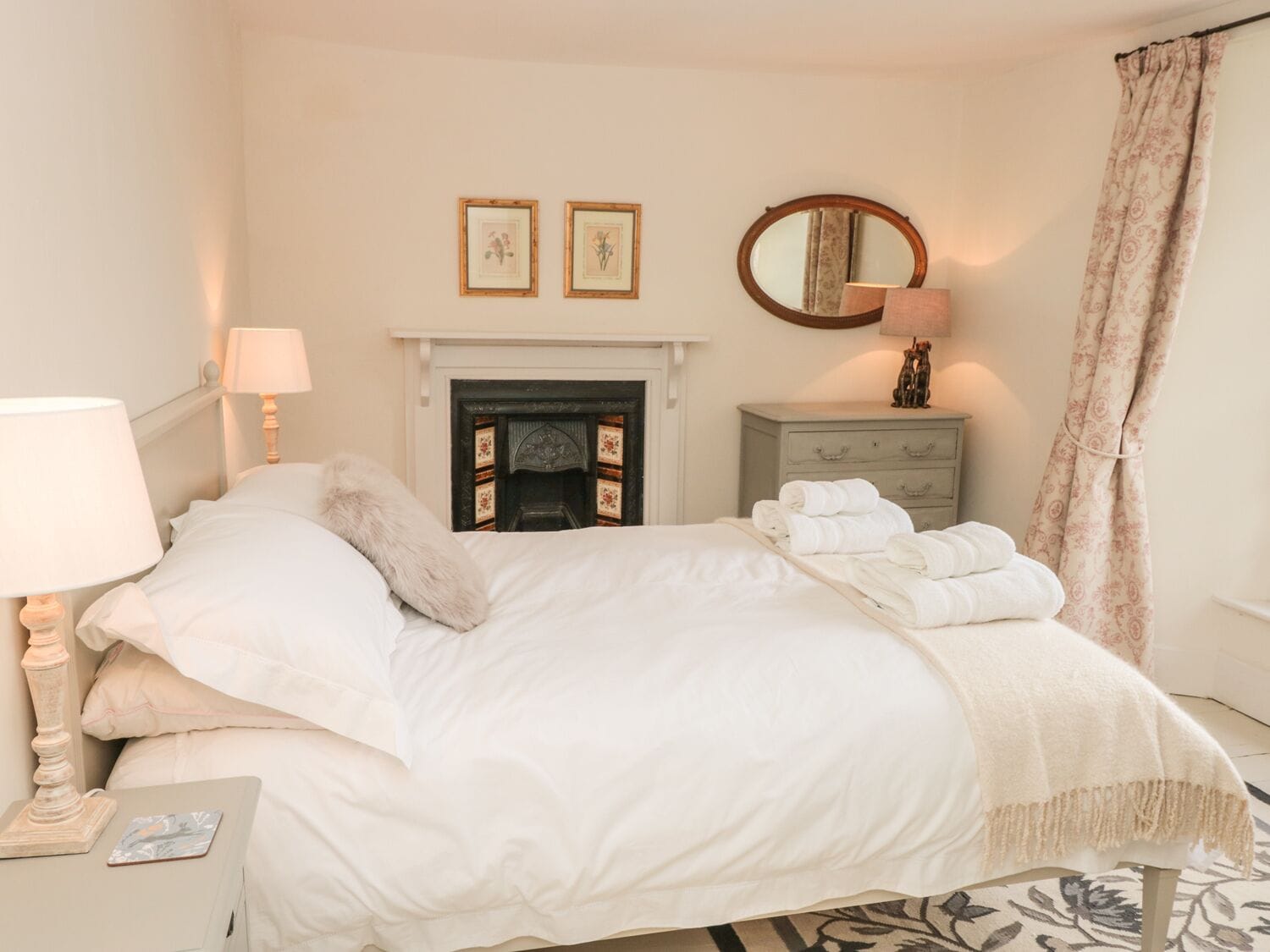 A bedroom with a bed, a bedside lamp, a dresser, a mirror, and a fireplace.