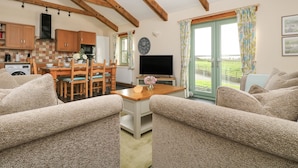 A cozy living room with a dining area, wooden beams, a TV, and a view of the countryside.