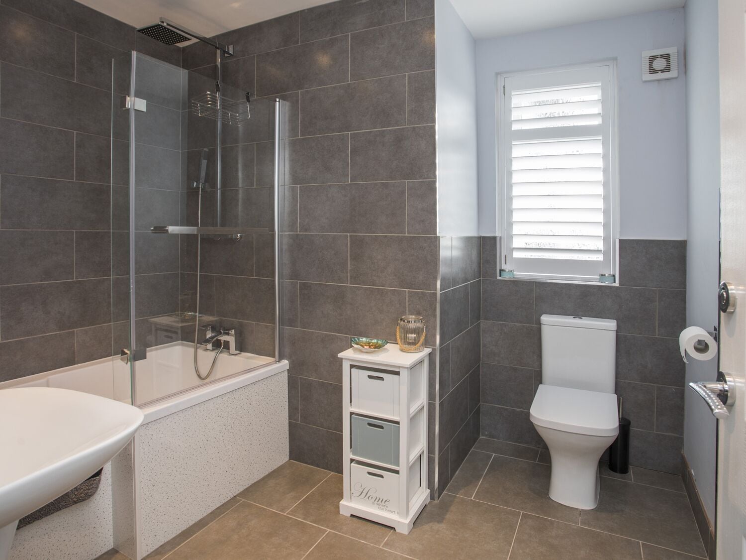 A modern bathroom with a glass-enclosed shower, a white bathtub, a toilet, and a small cabinet with a glass jar.