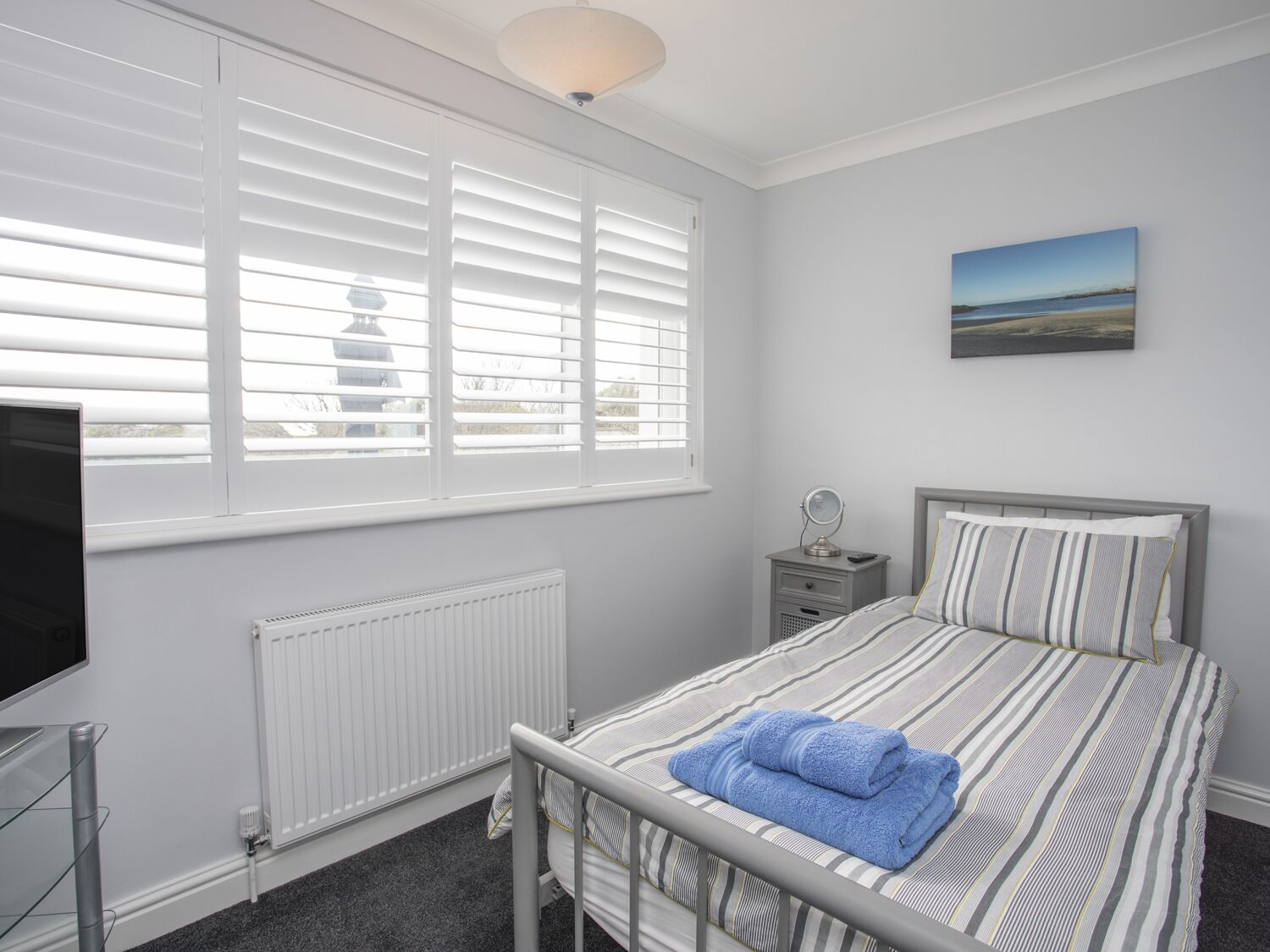 A bedroom with a bed, a bedside table with a lamp, a radiator, a TV, and a window with blinds.