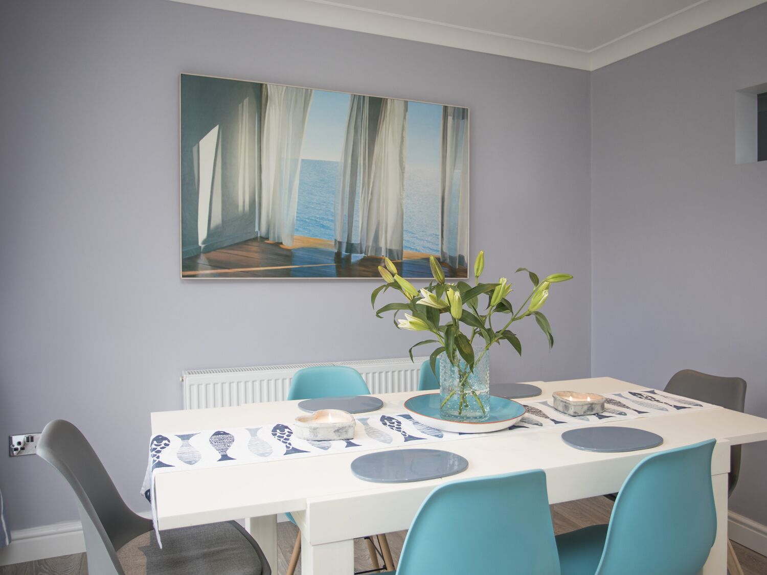 Un coin repas avec une table blanche, des chaises bleues, un vase de fleurs et un tableau représentant un paysage côtier.
