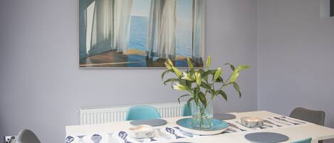 Un comedor con una mesa blanca, sillas azules, un florero con flores y un cuadro de una escena costera.