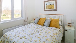 A bedroom with a bed, a window, a radiator, a bedside table, and a framed picture on the wall. - Beech Lodge (Newton Abbot)