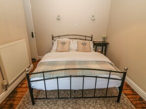 A neatly made bed with a metal frame, a bedside table with a vase, and a wall-mounted light fixture. - Glasfryn (Holyhead)