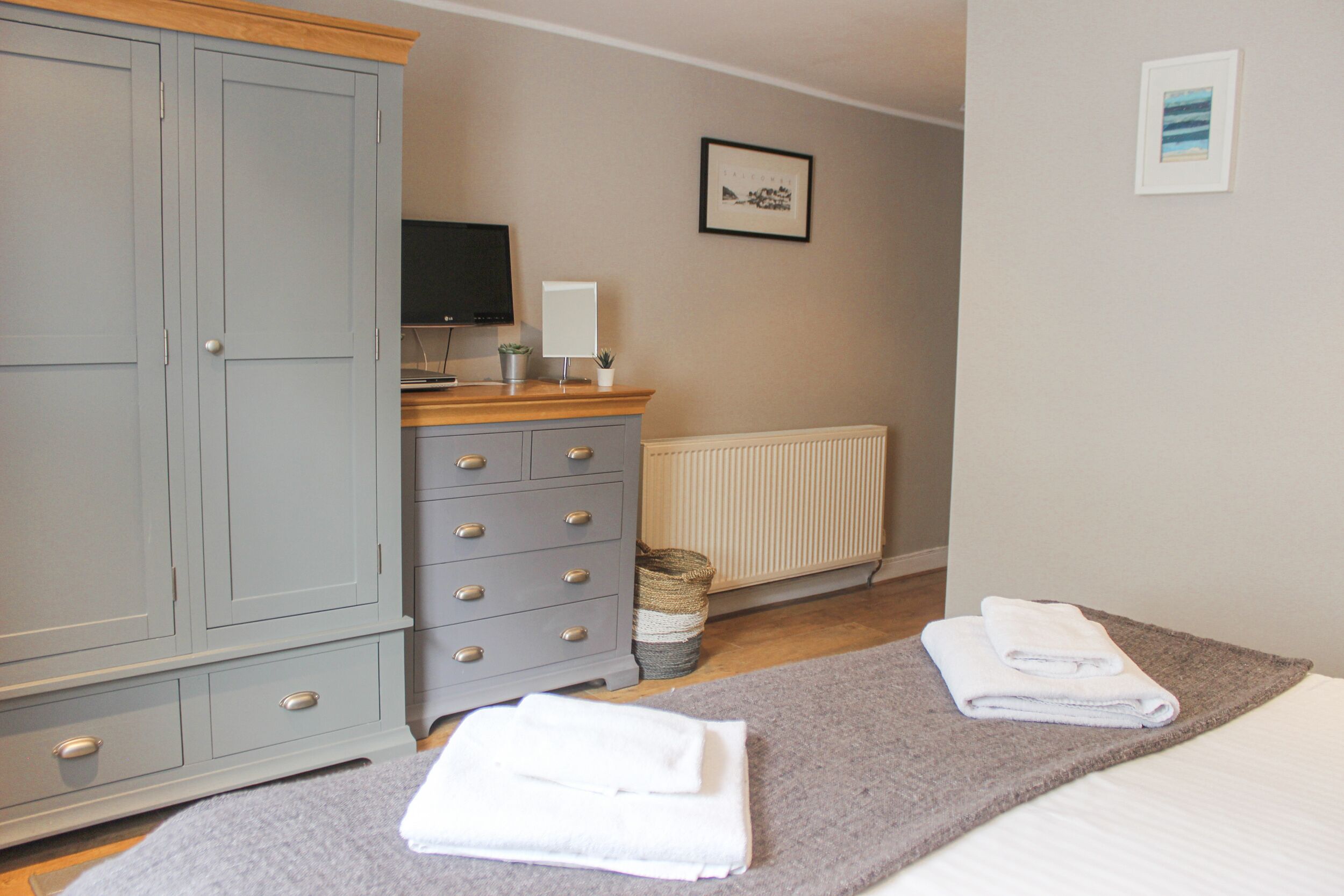 A bedroom with a grey wardrobe, a wooden dresser, a radiator, a framed picture on the wall, and two folded towels on a grey rug.