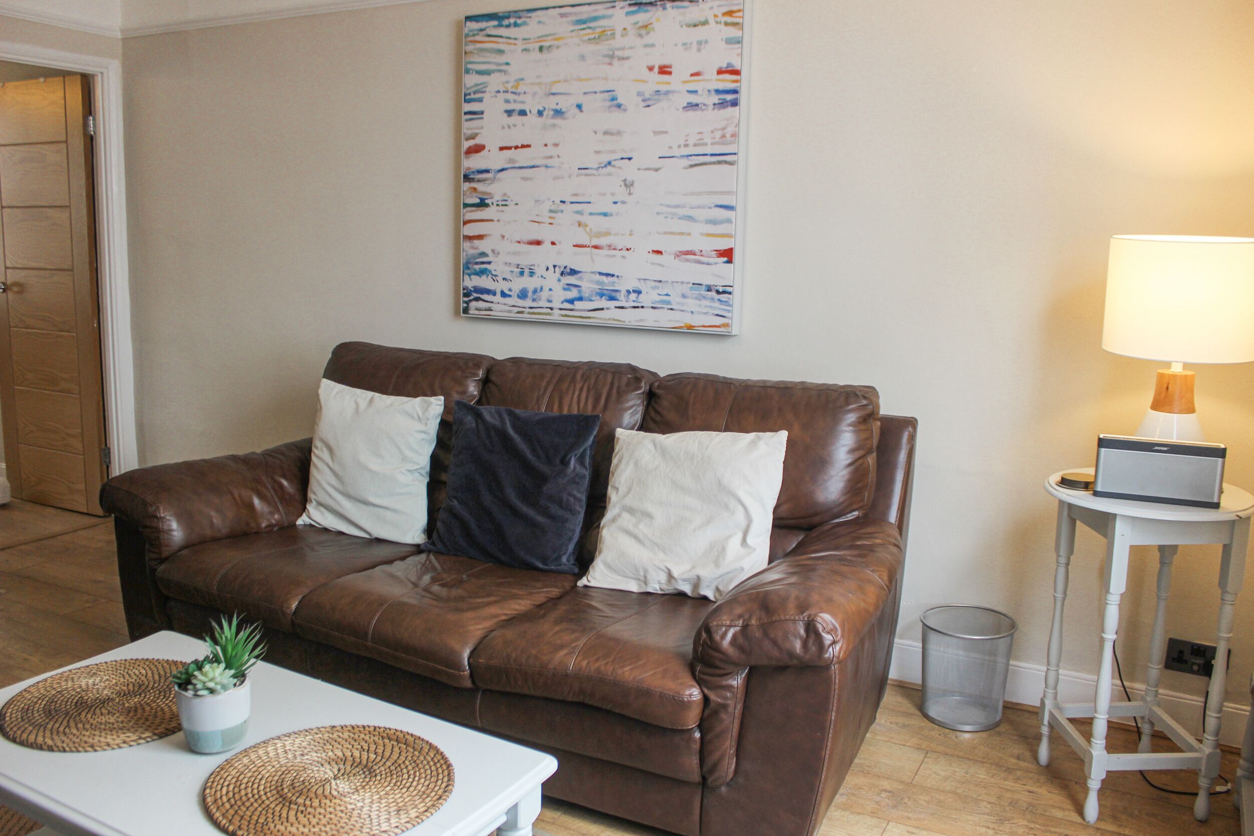 A brown leather sofa with white and dark cushions, a small table with a potted plant, a lamp on a side table, and a colorful abstract painting on the wall.
