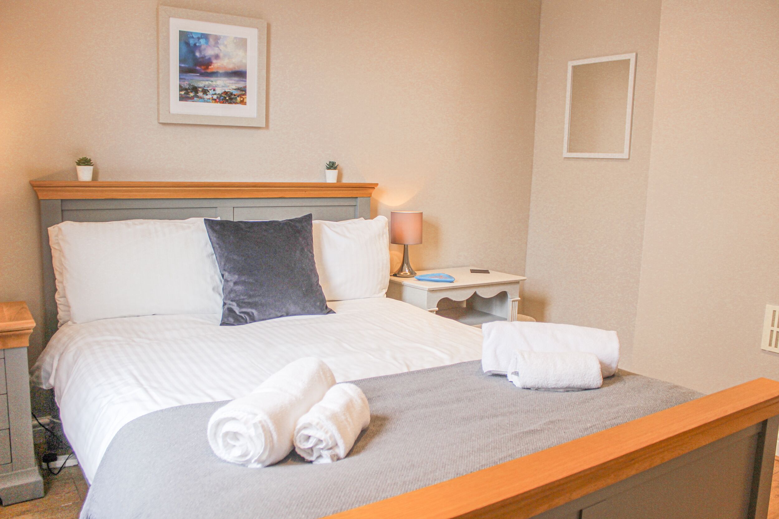 Una cama bien hecha con ropa de cama blanca y gris, un cabecero de madera y una mesita de noche con una lámpara y una pequeña planta.