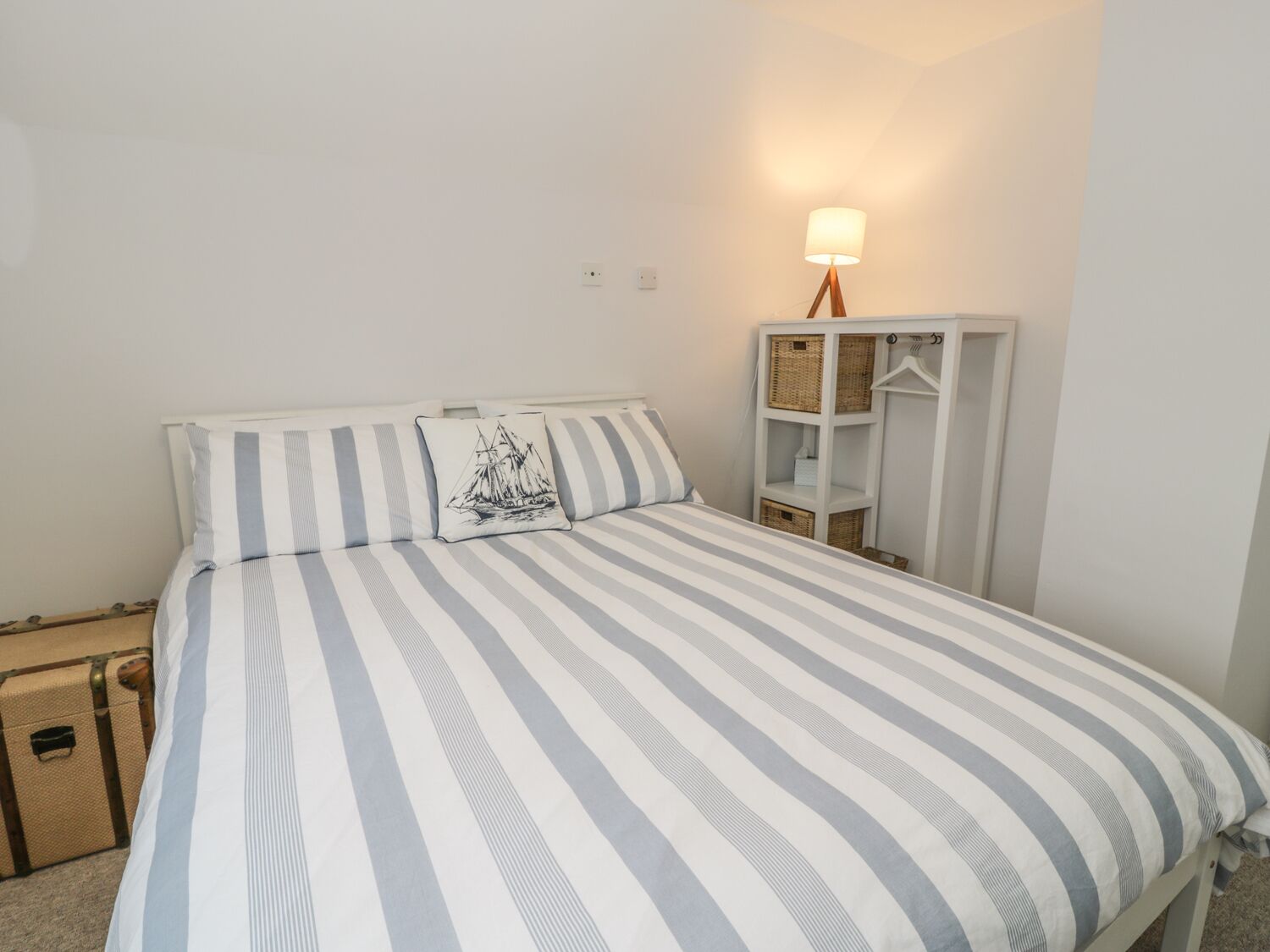 Quarto com colcha listrada, travesseiro com desenho de veleiro, luminária de mesa e guarda-roupa branco com prateleiras.