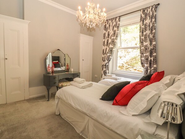 A bedroom with a bed, a dresser, a mirror, and a chandelier. - Beaulieu House (Llandudno)