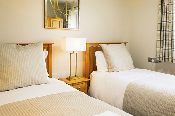 A hotel room with two beds, a nightstand with a lamp, a mirror, and a window with curtains.