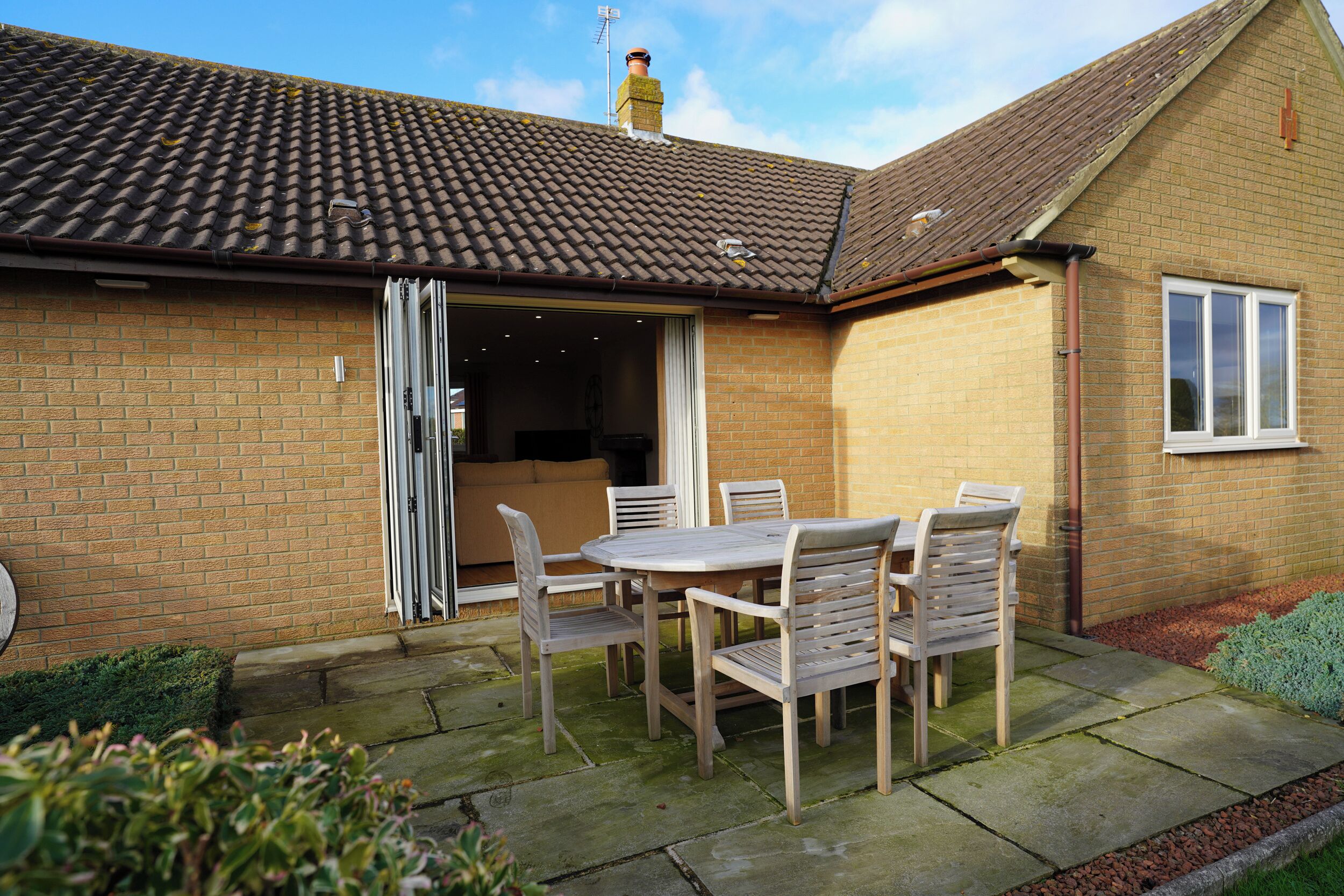 A brick house with a tiled roof, an open door leading to a room, and a patio with a table and chairs.
