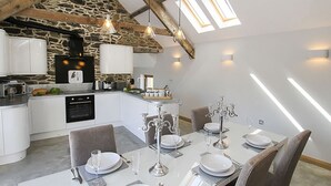 Una cuina moderna amb parets de pedra, zona de dinar amb taula preparada per a quatre persones, i un sostre d'estil loft amb bigues i finestres de sostre a la vista.
