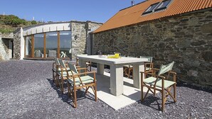 Edifici de pedra amb teulada a dues aigües, taula de formigó amb caires i una porta de vidre que dona a una habitació.