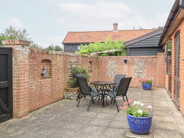 A patio area with a brick wall, a stone wall with a small archway, a round table with chairs, and a potted plant. - Lowbrook Cottage (Diss)