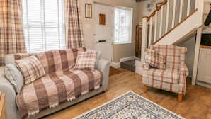 A cozy living room with a sofa and armchair, wooden beams on the ceiling, a staircase, and a patterned rug.