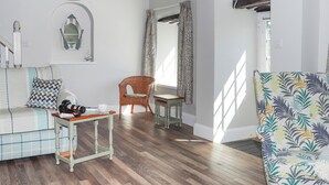 Un salon chaleureux avec un canapé, un fauteuil, une petite table et un fauteuil à motifs. On y trouve un escalier avec une rampe, une étagère murale agrémentée d’objets décoratifs et une fenêtre avec des rideaux.