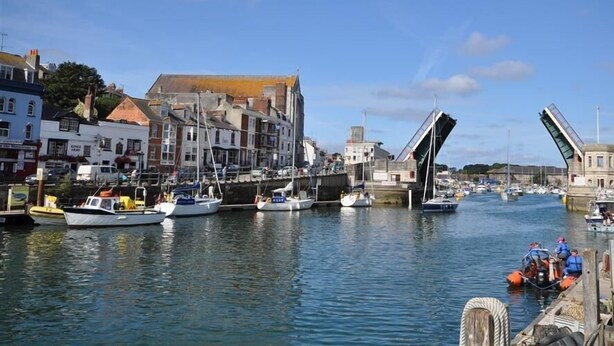 A harbor with boats docked, a bridge, and buildings along the waterfront. - Ocean Swell (Weymouth)