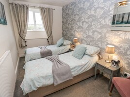 A bedroom with two beds, a window with curtains, a nightstand with a lamp, and a wall with a tree pattern.