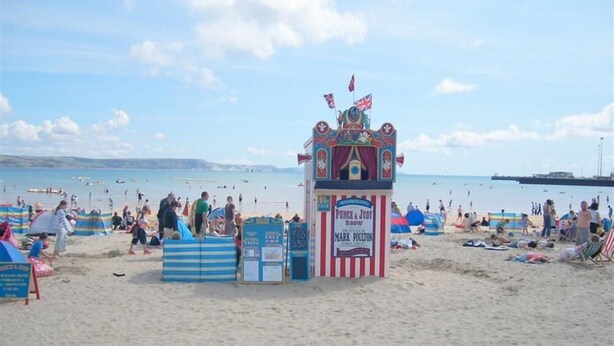 En strand med ett tält för en dockteaterföreställning av Punch och Judy, strandbesökare och en pir i bakgrunden.