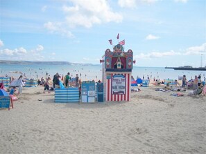 Ein Strand mit einem Zelt für eine Punch & Judy Vorstellung, Strandbesuchern und einem Steg im Hintergrund.