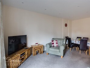 A living room with a TV, a sofa, a dining table with chairs, and a small side table with a bowl and a remote control. - Old Boathouse (Weymouth)