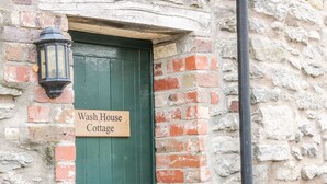 En träskylt med texten ”Wash House Cottage” sitter fast på en grön dörr, och ovanför dörren hänger en lykta. Allt detta är placerat framför en stenmur.