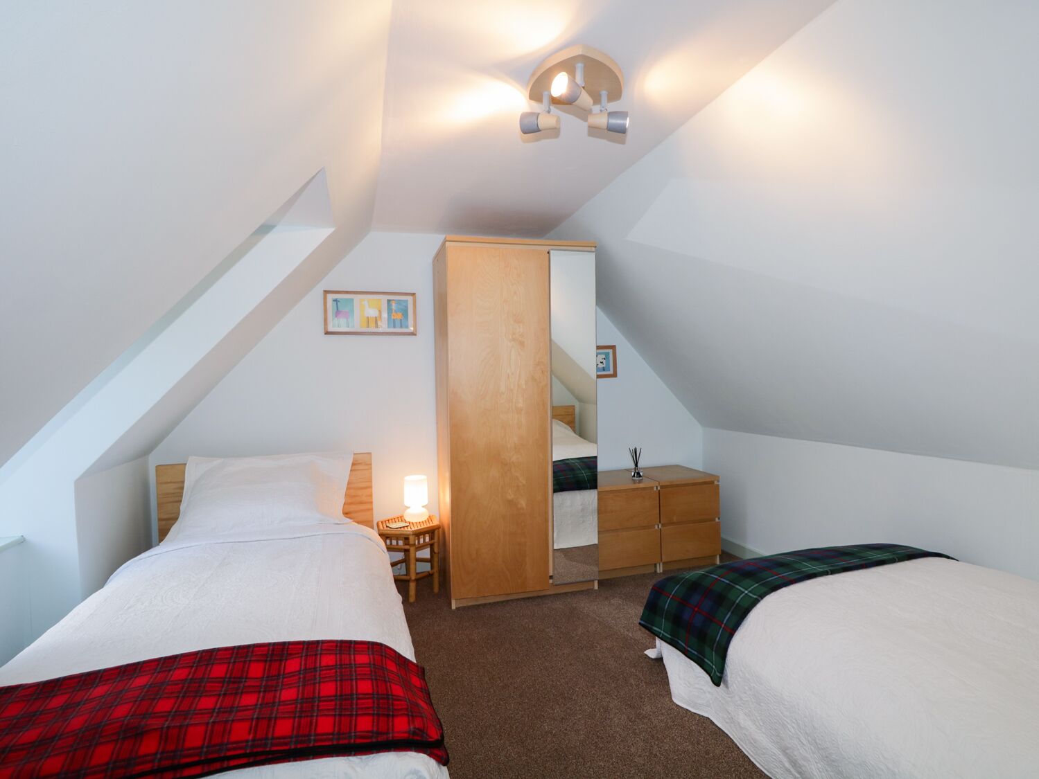 Une petite chambre mansardée, bien éclairée, avec deux lits, une commode en bois et un miroir.