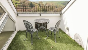 Eine kleine Terrasse mit Tisch und zwei Stühlen, Blick auf die Landschaft und eine weiße Wand.