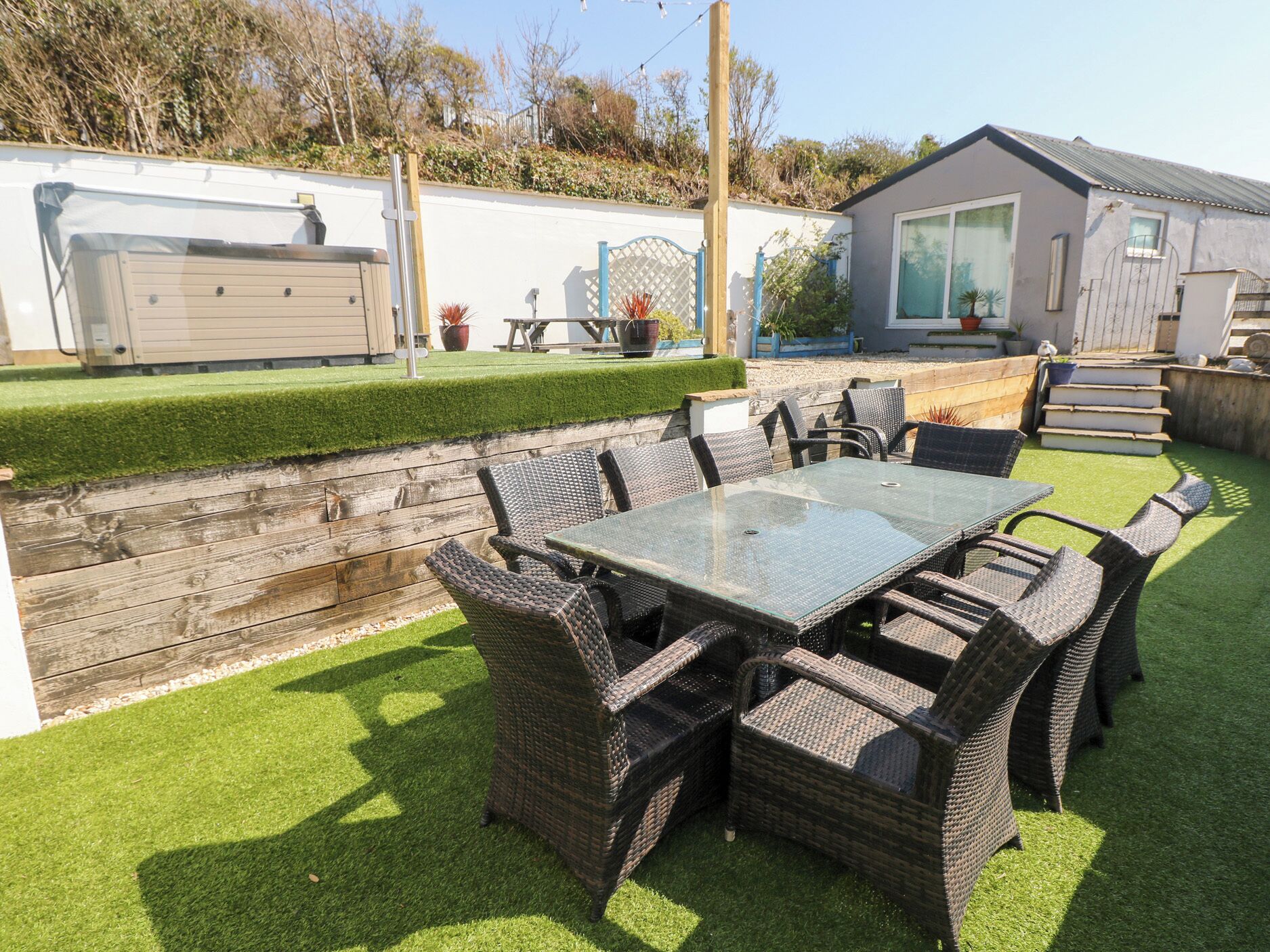 A patio area with a glass-top table and wicker chairs, surrounded by artificial grass and a wooden fence.