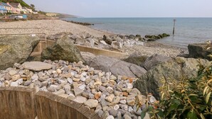 En stenstrand med en träpromenad som leder ner till havet, hus i bakgrunden och en livboj i vattnet.