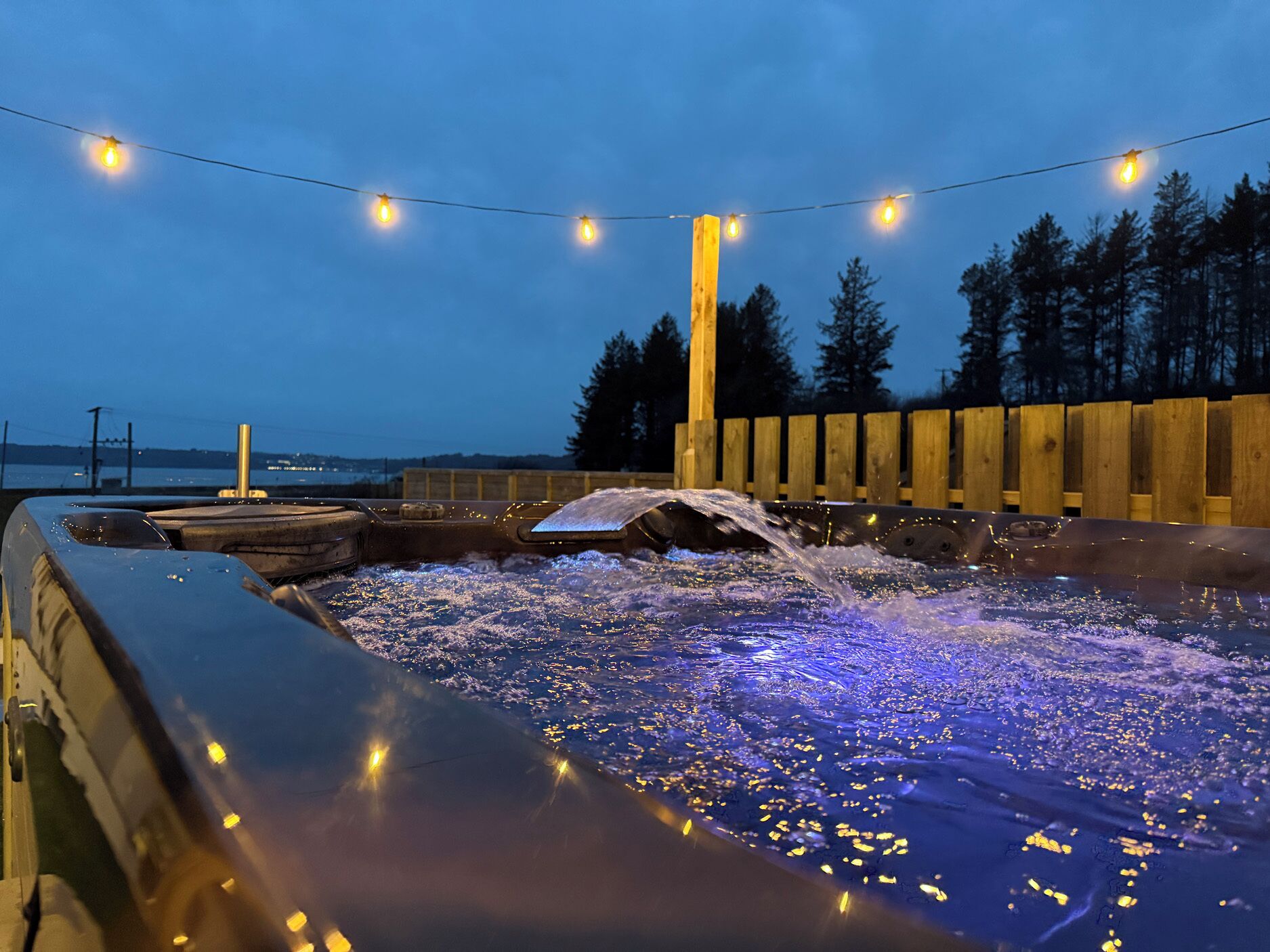 Un jacuzzi con chorros de agua, rodeado de cerdas de madera y luces de cuerda, con vistas a árboles y una masa de agua distante.