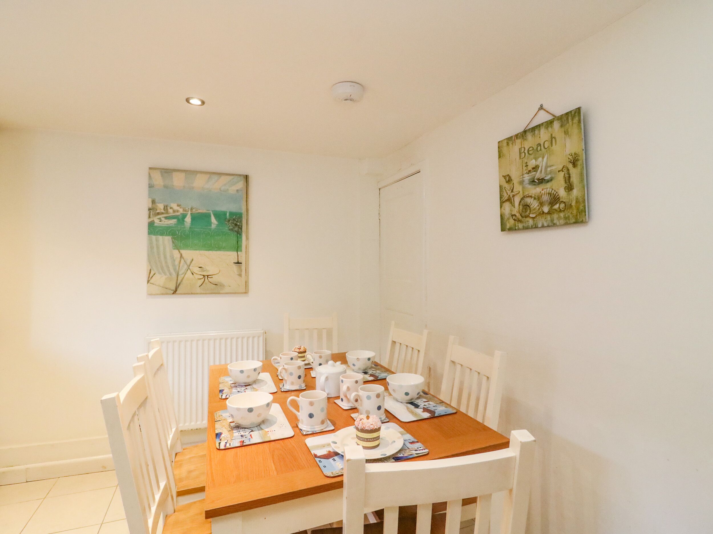 A dining area with a wooden table set for four, white chairs, and a beach-themed painting on the wall.