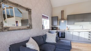 A modern living room with a grey sectional sofa, a large ornate mirror, and a kitchen with grey cabinets and a microwave. - The Stables (Scarborough)