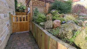 A wooden gate leading to a garden with a pergola, a brick pathway, and a stone wall. - The Stables (Scarborough)
