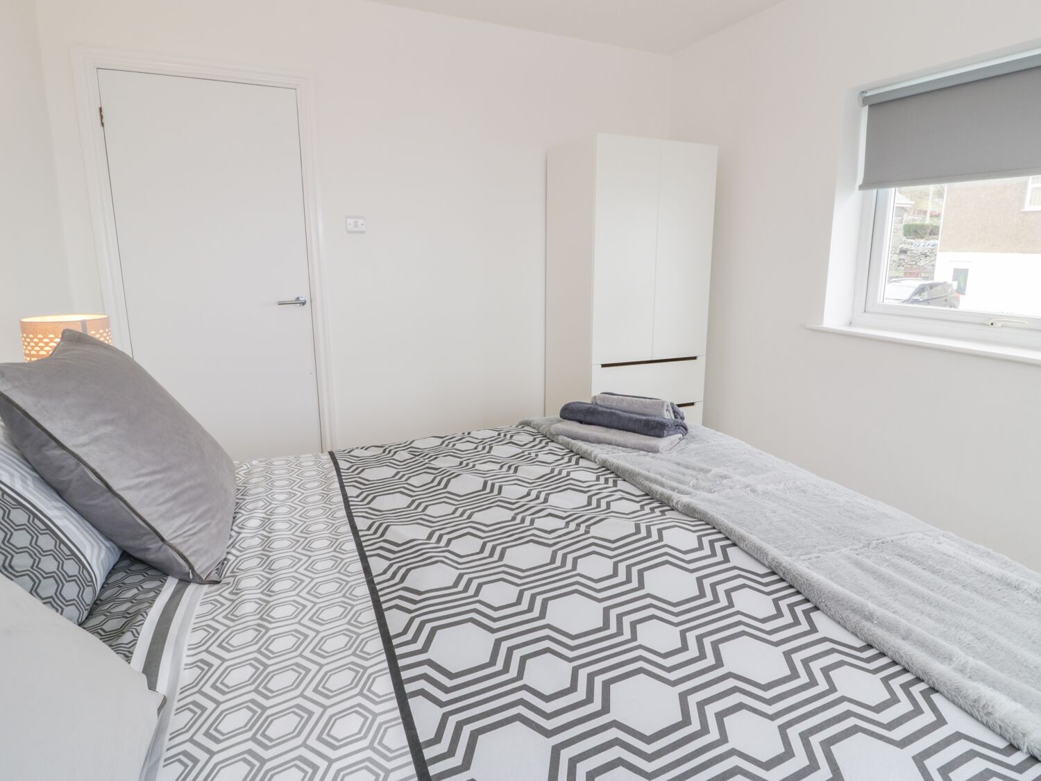 Ein kleines Zimmer mit einem Einzelbett, einer geometrisch gemusterten Tagesdecke, einem weißen Kleiderschrank und einem Fenster mit Blick nach außen.