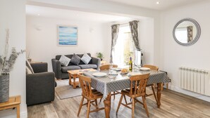 Un coin repas avec une table dressée pour quatre personnes, entourée de chaises en bois. On aperçoit à l’arrière-plan un canapé gris avec des coussins, une plante en pot et un miroir fixé au mur.