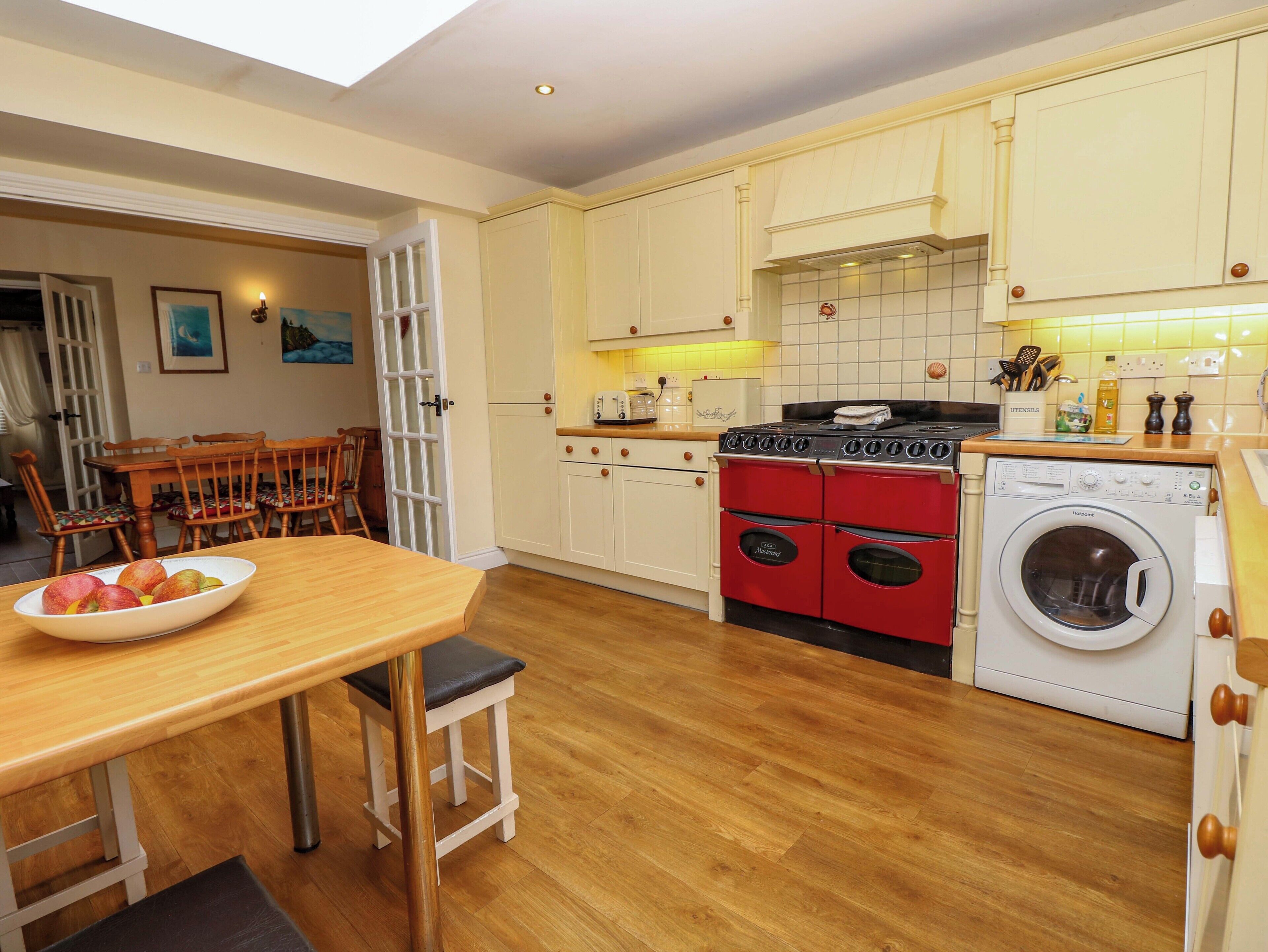 A compact kitchen with a dining area, featuring a wooden table, chairs, a red stove, a washing machine, and a tiled backsplash.
