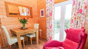 A wooden cabin interior with a dining table, chairs, a red leather chair, a mirror, and a window with floral curtains. - Borah Lodge (Penzance)